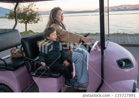 Mother And Child Riding A Pink Electric Vehicle By The Lake Shore. Mother And Child Riding A Pink Electric Vehicle By The Lake Shore. 135727891