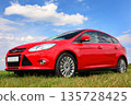 Red car on a blue sky background with white clouds. The car is standing in the field on the green grass. 135728425