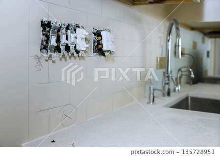 Workers are installing electrical outlets on wall of kitchen, wall has white tiles, sink faucet are visible nearby. Workers are installing electrical outlets on wall of kitchen, wall has white tiles, sink faucet are visible nearby. 135728951
