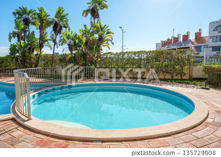A bright and inviting pool area features clear water and lush tropical plants under a clear blue sky, perfect for relaxing on a sunny day 135729008