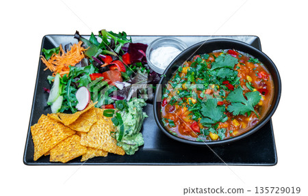Colorful vegetarian chili served in a bowl with fresh salad, tortilla chips, and guacamole on a black plate, showcasing a vibrant meal presentation 135729019