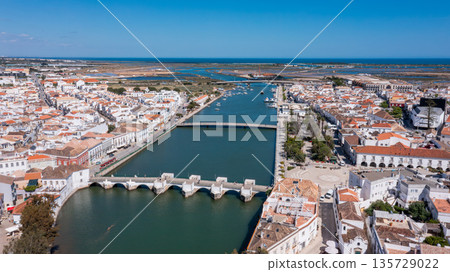 The scene shows Tavira in Algarve from above, highlighting the Gilao river, several Roman bridge, and the town's layout under clear blue skies in Portugal The scene shows Tavira in Algarve from above, highlighting the Gilao river, several Roman bridge, and the town's layout under clear blue skies in Portugal 135729022