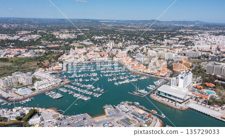 Aerial view of Vilamoura marina in Portugal showing numerous boats docked in the water and buildings along the shore under a clear blue sky. Aerial Aerial view of Vilamoura marina in Portugal showing numerous boats docked in the water and buildings along the shore under a clear blue sky. Aerial 135729033