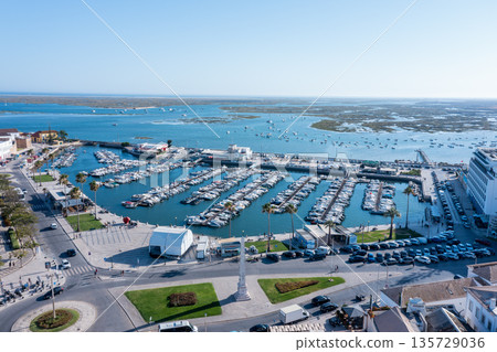 People visit the marina in Faro, Portugal with many boats docked and Ria Formosa in the distance. The sun shines brightly over the old town area 135729036