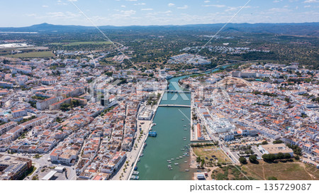 Tavira in Algarve is shown from above, with houses lined along the Gilao river and boats docked nearby. The scene highlights the city layout and natural features Tavira in Algarve is shown from above, with houses lined along the Gilao river and boats docked nearby. The scene highlights the city layout and natural features 135729087