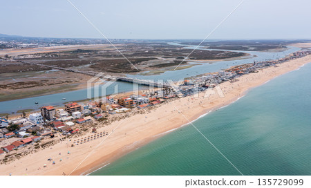 A clear aerial view of Praia de Faro shows the beach, water, and nearby structures along the shoreline in Ria Formosa, Portugal. People enjoy the sunny day 135729099