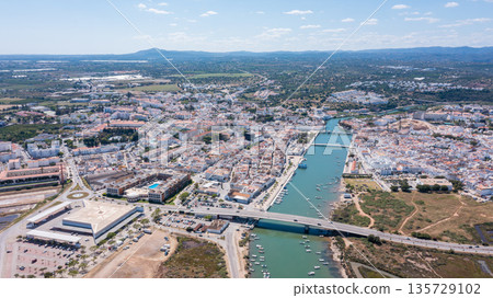 Tavira, a town in Algarve, is seen from the sky with waterways, buildings, and fields. Boats float in the river under bright sunlight and blue sky 135729102