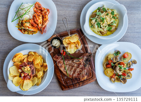 Delicious assortment of gourmet dishes featuring grilled shrimp, steak, pasta, and crispy chips arranged beautifully on a wooden table Delicious assortment of gourmet dishes featuring grilled shrimp, steak, pasta, and crispy chips arranged beautifully on a wooden table 135729111