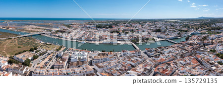 Tavira in Algarve is seen from above, featuring the river and white buildings with a clear sky. The view captures the town's layout and surrounding landscape. Panorama River Gilao and Roman bridge Tavira in Algarve is seen from above, featuring the river and white buildings with a clear sky. The view captures the town's layout and surrounding landscape. Panorama River Gilao and Roman bridge 135729135