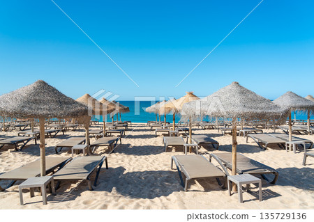 Sun loungers are arranged in rows under thatched umbrellas on a sandy beach. The sea meets the clear blue sky in the background on a sunny day Sun loungers are arranged in rows under thatched umbrellas on a sandy beach. The sea meets the clear blue sky in the background on a sunny day 135729136