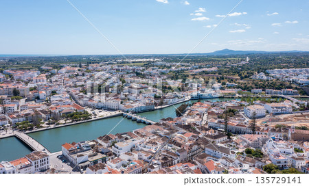 Tavira in Algarve is captured from above, showing its buildings, Gilao river, and green areas under a clear sky with some clouds Tavira in Algarve is captured from above, showing its buildings, Gilao river, and green areas under a clear sky with some clouds 135729141
