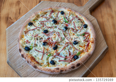Delicious pizza topped with ham, olives, and green peppers on a wooden board, showcasing a mouthwatering culinary delight for food lovers Delicious pizza topped with ham, olives, and green peppers on a wooden board, showcasing a mouthwatering culinary delight for food lovers 135729169