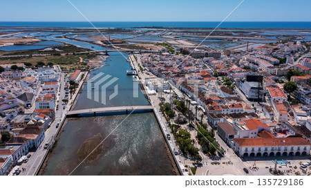 Aerial view of Tavira with modern bridge over the Gilao River, traditional buildings, salt flats and Atlantic Ocean in the background under clear blue sky in Algarve, Portugal.  135729186