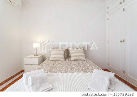 A well-lit bedroom features a double bed with striped pillows and freshly arranged towels. A bedside table holds a lamp, against crisp white walls and a neat closet A well-lit bedroom features a double bed with striped pillows and freshly arranged towels. A bedside table holds a lamp, against crisp white walls and a neat closet 135729188
