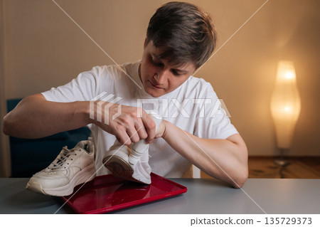 Portrait of young adult man applying white protective paint to dirty sneaker, providing shoe care, renewal and restoration, cleaning footwear and demonstrating maintenance process. Portrait of young adult man applying white protective paint to dirty sneaker, providing shoe care, renewal and restoration, cleaning footwear and demonstrating maintenance process. 135729373