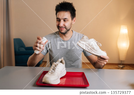 Portrait of positive young man holding white sneaker and shoe cleaner bottle, preparing to clean footwear at home, demonstrating process of refreshing shoes and maintaining appearance. Portrait of positive young man holding white sneaker and shoe cleaner bottle, preparing to clean footwear at home, demonstrating process of refreshing shoes and maintaining appearance. 135729377