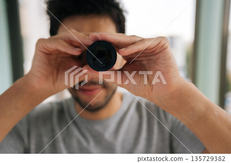 Close-up portrait of bearded man curiously peering out window using monocular, watching with emotion. Engaging in voyeurism. Observing surveillance. Peeping through curtains. Maintaining vigilance. 135729382
