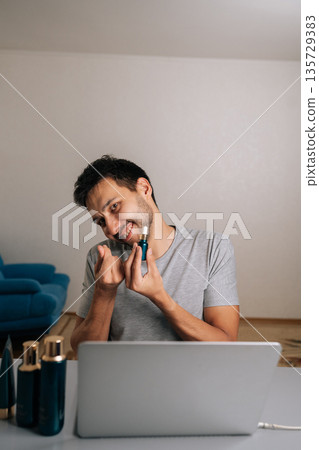 Vertical portrait of male vlogger creating skincare content online, reviewing beauty serum and making heart gesture with hands, connecting with audience about personal care, cosmetic products. 135729383
