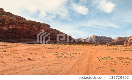 Rocky massifs on red sand desert, bright cloudy sky in background, small  vehicle and camel at distance - typical scenery in Wadi Rum, Jordan 135729892