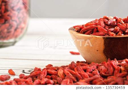 Dried goji berries in wooden bowl, scattered over white boards table under, blurred large glass jar full of fruits in background Dried goji berries in wooden bowl, scattered over white boards table under, blurred large glass jar full of fruits in background 135730459