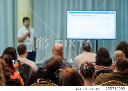 Presenter speaks at conference to attentive audience with large screen and notes during professional seminar Presenter speaks at conference to attentive audience with large screen and notes during professional seminar 135730903
