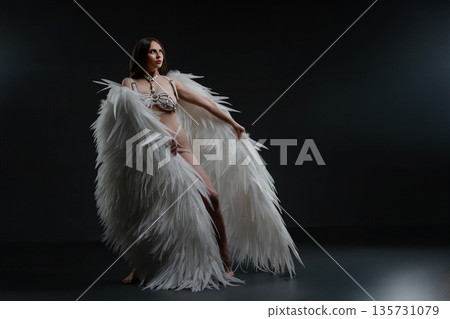 Elegant woman in white angel wings and harness poses dramatically in a dark studio. 135731079