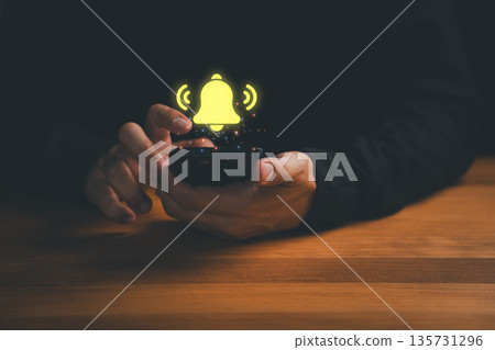 Close-up of hands holding a smartphone with glowing notification bell icon above the screen. Concept for mobile alert, app notification, message reminder, and digital communication signal. Close-up of hands holding a smartphone with glowing notification bell icon above the screen. Concept for mobile alert, app notification, message reminder, and digital communication signal. 135731296