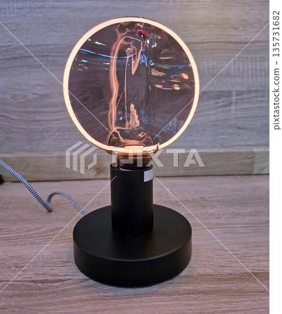 A decorative lamp sits on a wooden table in a modern room. The glass bulb glows warmly, creating a soft light that fills the space. It adds charm to the surroundings during the evening. 135731682