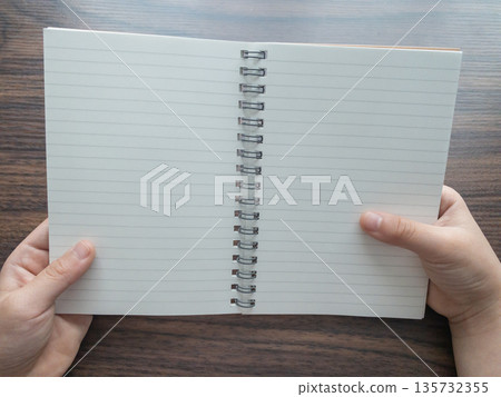 孩子雙手拿著空白筆記本,坐在木桌旁。 孩子雙手拿著空白筆記本,坐在木桌旁。 135732355