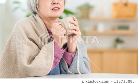一位老婦人在客廳裡感到寒冷,肩上披著毯子(熱飲,取暖,取暖) 一位老婦人在客廳裡感到寒冷,肩上披著毯子(熱飲,取暖,取暖) 135733093