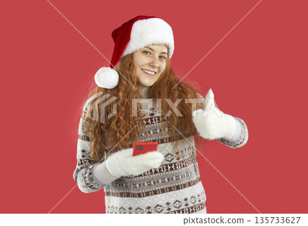 Pretty redhead Christmas girl in Santa hat showing thumb up holding smartphone on red background. 135733627