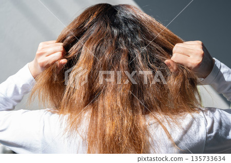 Rear view of young woman while touching her messy and frizzy hair. Hair standing up or curling away from the main hair, creating a fuzzy, unruly, or rough texture, often due to dryness or humidity. 135733634