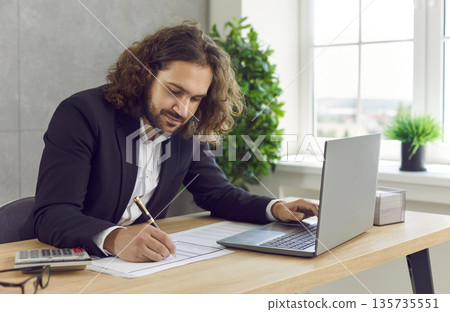 Male accountant working with business documents and using laptop at desk in his office Male accountant working with business documents and using laptop at desk in his office 135735551