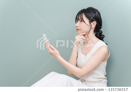 一位年輕的亞洲女性在家中看著智慧型手機,若有所思(擔憂、嚴肅、思考、計劃) 一位年輕的亞洲女性在家中看著智慧型手機,若有所思(擔憂、嚴肅、思考、計劃) 135736557