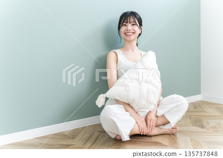 一位面帶微笑的年輕亞洲女性在家中客廳抱著靠墊放鬆休息。 一位面帶微笑的年輕亞洲女性在家中客廳抱著靠墊放鬆休息。 135737848