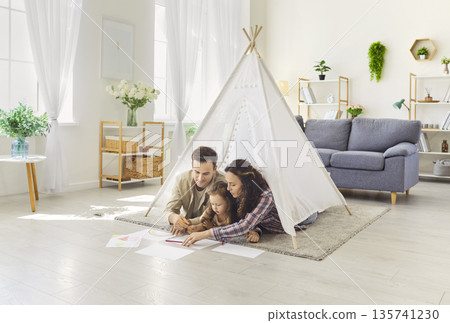 Happy parents and daughter drawing together on floor at home in cozy play tent Happy parents and daughter drawing together on floor at home in cozy play tent 135741230