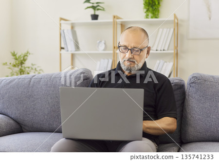 Serious senior man wearing glasses using laptop on comfortable sofa at home Serious senior man wearing glasses using laptop on comfortable sofa at home 135741233
