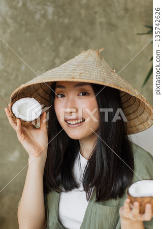 A young Asian woman wearing a traditional conical hat smiles while holding a halved coconut in each hand, showcasing tropical fruit A young Asian woman wearing a traditional conical hat smiles while holding a halved coconut in each hand, showcasing tropical fruit 135742626