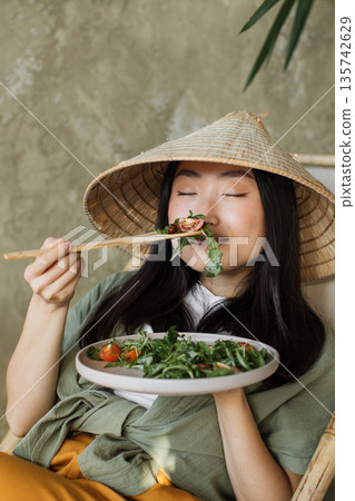 A young woman wearing a traditional conical hat enjoys a healthy salad with cherry tomatoes and greens, using chopsticks to eat A young woman wearing a traditional conical hat enjoys a healthy salad with cherry tomatoes and greens, using chopsticks to eat 135742629