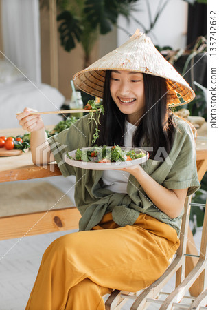 A young Asian woman wearing a conical hat enjoys a healthy salad with chopsticks at a wooden table, surrounded by greenery A young Asian woman wearing a conical hat enjoys a healthy salad with chopsticks at a wooden table, surrounded by greenery 135742642