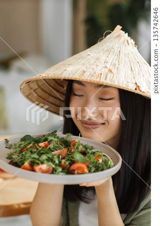 A young woman wearing a traditional conical hat savors the aroma of a vibrant, healthy salad filled with greens and tomatoes A young woman wearing a traditional conical hat savors the aroma of a vibrant, healthy salad filled with greens and tomatoes 135742646