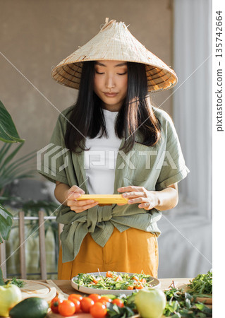 A young Asian woman wearing a conical hat takes a picture of a fresh salad on her phone while preparing food in a bright, natural setting A young Asian woman wearing a conical hat takes a picture of a fresh salad on her phone while preparing food in a bright, natural setting 135742664