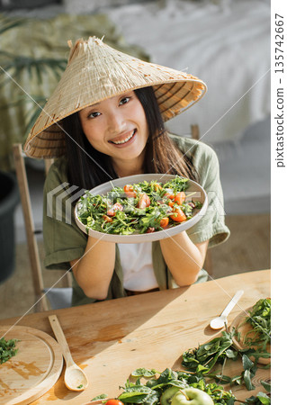 A smiling young Asian woman wearing a conical hat holds a fresh salad bowl while sitting at a wooden table with ingredients 135742667