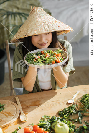 A young woman wearing a conical hat savors the aroma of a freshly prepared salad filled with greens and tomatoes 135742668