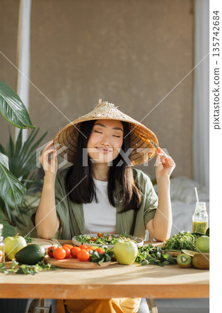 A serene Asian woman wearing a conical hat smiles with her eyes closed while surrounded by fresh fruits and vegetables, embodying a healthy lifestyle A serene Asian woman wearing a conical hat smiles with her eyes closed while surrounded by fresh fruits and vegetables, embodying a healthy lifestyle 135742684
