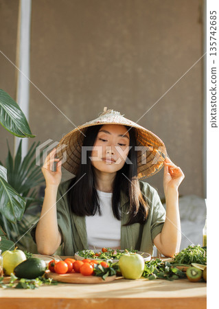 A young Asian woman wearing a traditional conical hat sits at a table laden with fresh fruits and vegetables, preparing a healthy meal A young Asian woman wearing a traditional conical hat sits at a table laden with fresh fruits and vegetables, preparing a healthy meal 135742685