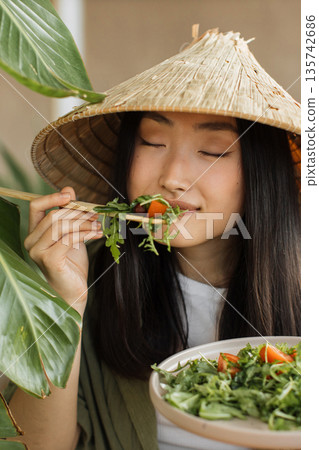 A young Asian woman wearing a conical hat enjoys a fresh salad with chopsticks, surrounded by lush green foliage 135742686