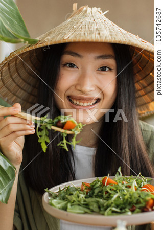 A young Asian woman wearing a conical hat smiles while eating a fresh green salad with cherry tomatoes using chopsticks A young Asian woman wearing a conical hat smiles while eating a fresh green salad with cherry tomatoes using chopsticks 135742687