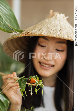 A young woman wearing a conical hat enjoys a fresh salad with cherry tomatoes and greens, highlighting healthy eating and Asian culture 135742689