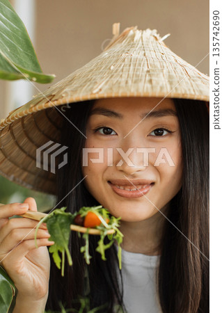 A young Asian woman wearing a conical hat enjoys a healthy salad with a wooden spoon, showcasing fresh greens and a cherry tomato 135742690